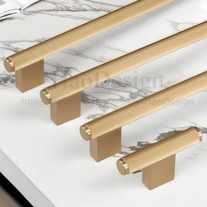 May include: Close-up of several gold-colored cabinet pulls with a ribbed design. The pulls are rectangular with a cylindrical handle. The pulls are on a white marble surface. The Etsy shop name and URL are visible in the image.