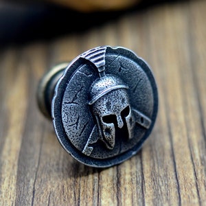 May include: A silver-toned metal cufflink featuring a detailed design of a Spartan helmet with a shield.