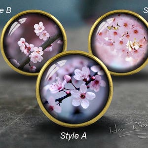 Peut inclure: Trois cabochons ronds en verre décoratifs, de couleur dorée, avec des images différentes de fleurs de cerisier roses. Les cabochons sont étiquetés "Style A", "Style B" et "Style C".