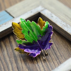 May include: A gold-toned brooch in the shape of a maple leaf. The leaf is layered with green, purple, and gold enamel.