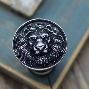 May include: A round knob made of silver-toned metal, featuring a detailed lion's head design. The lion's mane is textured and the face is detailed with a fierce expression.