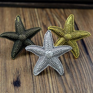 Puede incluir: Tres estrellas de mar decorativas en diferentes acabados: plata, oro y bronce. Cada estrella de mar tiene una superficie texturizada con pequeños puntos en relieve.