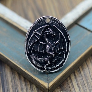 Peut inclure: Un pendentif argenté avec un motif de dragon détaillé. Le pendentif est de forme ovale et a une surface texturée.