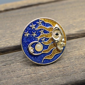 Peut inclure: Une épingle dorée avec un motif bleu et or. Le motif représente un soleil stylisé avec un visage et une lune croissante avec un centre perlé. L'arrière-plan est un émail bleu avec des paillettes dorées et des étoiles.
