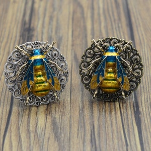 May include: Two silver and gold bee brooches with blue and yellow enamel. The bees are surrounded by a filigree design.