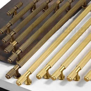 May include: A collection of brass cabinet pulls and handles in various sizes. The hardware features a ribbed cylindrical design and a warm, golden finish. The pulls are arranged on a white and brown surface.