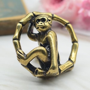 Puede incluir: Un amuleto de mono dorado con un dise&ntilde;o detallado, sentado dentro de un marco de bamb&uacute; circular. El amuleto es un animal del zod&iacute;aco chino tradicional.