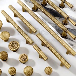May include: A collection of antique brass cabinet hardware, including various sizes of knobs and pulls. The hardware features a hammered texture and a warm, golden hue. The arrangement includes both round knobs and bar-style pulls.