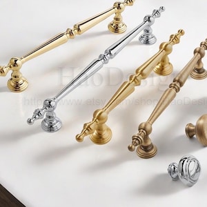 Peut inclure: Une collection de quincaillerie décorative pour meubles, en finitions or, argent et bronze. L'assortiment comprend plusieurs poignées et boutons, avec des motifs ornés et un aspect poli. La quincaillerie est présentée sur une surface blanche.