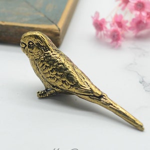 Peut inclure: Figurine de perruche en métal doré. Le petit oiseau présente des détails de plumes complexes et une longue queue. La figurine est posée sur un fond blanc avec le texte "Hao Design" et une adresse de site web.