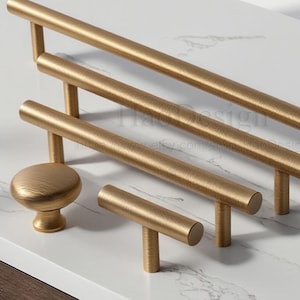 May include: A selection of gold-coloured cabinet hardware, including bar pulls and a knob, displayed on a white surface. The hardware features a textured finish and a modern design, suitable for various furniture styles.
