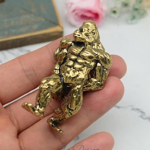 May include: A gold-coloured metal figurine of a gorilla sitting with its arms crossed. The figurine is detailed with muscle definition and has a textured surface.
