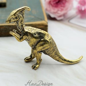 以下が含まれることがあります： 長い首と尾を持つ金色の金属製の恐竜の置物。恐竜は白い表面に立っています。置物は約12.5cmの高さです。