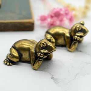 以下が含まれることがあります： 白い大理石の表面に、休息ポーズをとった小さな金色のカエルの置物が2つ置かれています。