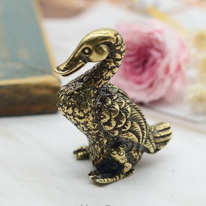 May include: A small, gold-coloured brass duck figurine with detailed feathers and a textured surface. The duck is sitting with its head turned to the side.