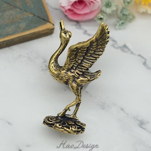 May include: A gold-toned metal crane figurine with its wings spread, standing on a cloud-shaped base. The crane is facing to the right and has a detailed design.