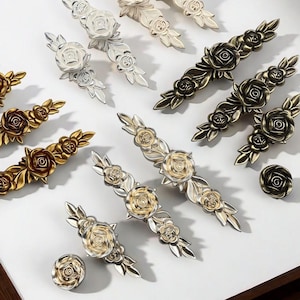 May include: An assortment of decorative cabinet hardware featuring rose designs. The hardware includes knobs and pulls in various metallic finishes, including gold, silver, and bronze. The rose details are intricately designed, adding a touch of elegance to any furniture piece.