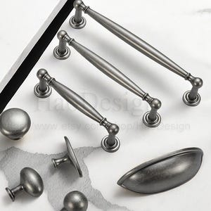 Puede incluir: Un surtido de herrajes de gabinete de estilo antiguo en un acabado plateado apagado. La colección incluye varios tiradores, pomos y un tirador de taza, todos dispuestos sobre una superficie blanca.