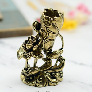 以下が含まれることがあります： 金色の金属製の蓮の花のデザインで、2匹の魚と花瓶があります。花瓶は蓮の花のつぼみの形をしています。