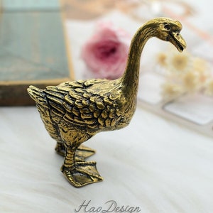 May include: A gold-colored metal goose figurine. The goose is standing with its head turned to the side. The figurine is detailed with feathers and has a realistic appearance.
