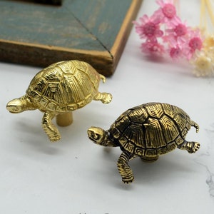 Könnte beinhalten: Zwei goldene Schubladengriffe in Form von Schildkröten. Die Schildkröten sind mit strukturierten Schalen verziert und haben eine leicht abgenutzte Oberfläche.