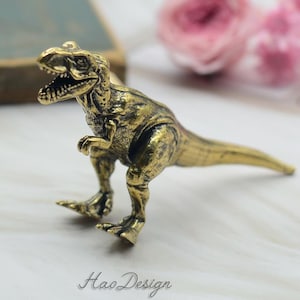 May include: A gold-coloured metal dinosaur figurine. The dinosaur is standing on all fours with its mouth open and its tail extended. The figurine is detailed with textured scales and sharp teeth.