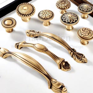 May include: A selection of antique brass cabinet knobs and handles. The knobs display intricate floral and geometric patterns. The handles are curved and detailed. The hardware is arranged on a white surface.