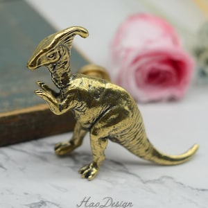 May include: A gold-colored dinosaur-shaped cabinet knob or drawer pull. The dinosaur is standing on all fours with its head tilted down. The dinosaur has a long tail and a crest on its head.