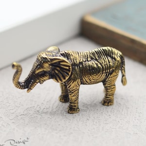 Puede incluir: Una pequeña figurita de elefante de latón de color dorado con una superficie texturizada. El elefante está de pie sobre una superficie gris claro.