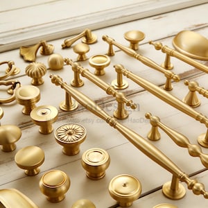 May include: A collection of gold-colored cabinet hardware, including knobs and pulls, arranged on a light-colored wooden surface. The hardware features various designs, from simple round knobs to ornate pulls with detailed handles. The image showcases a variety of styles and sizes.