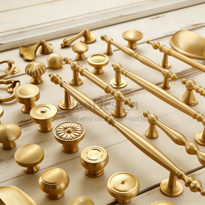 May include: A collection of gold-colored cabinet hardware, including knobs and pulls, arranged on a light-colored wooden surface. The hardware features various designs, from simple round knobs to ornate pulls with detailed handles. The image showcases a variety of styles and sizes.