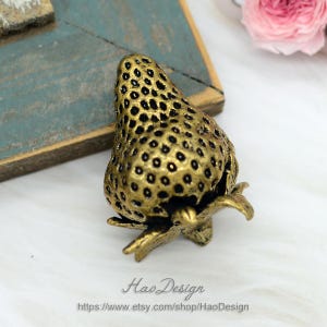 Op de afbeelding: Een gouden metalen ladegreep in de vorm van een aardbei. De greep heeft een getextureerd oppervlak en een steel met bladeren.