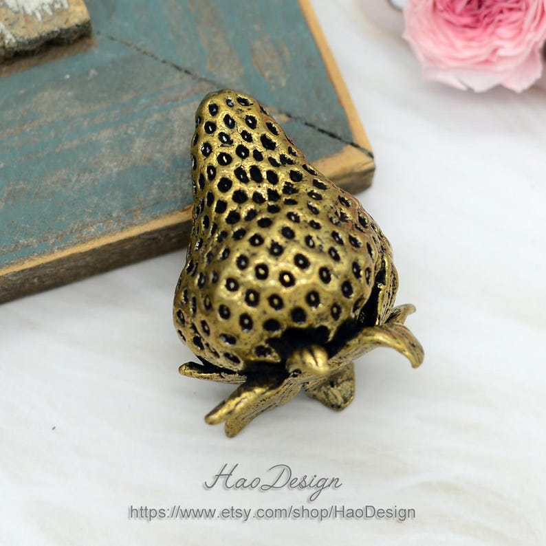 Op de afbeelding: Een gouden metalen ladegreep in de vorm van een aardbei. De greep heeft een getextureerd oppervlak en een steel met bladeren.