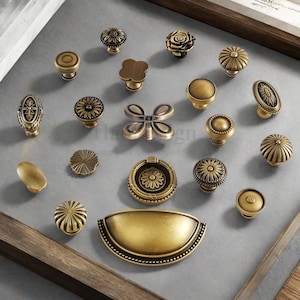 May include: A collection of antique brass cabinet knobs and pulls in various designs. The knobs include floral, geometric, and oval shapes. The hardware is arranged on a gray surface.