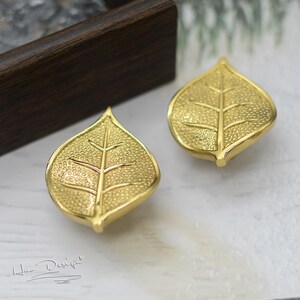 K&ouml;nnte beinhalten: Ein Paar goldene Ohrringe in Blattform. Die Bl&auml;tter haben eine strukturierte Oberfl&auml;che und ein detailliertes Adernmuster.