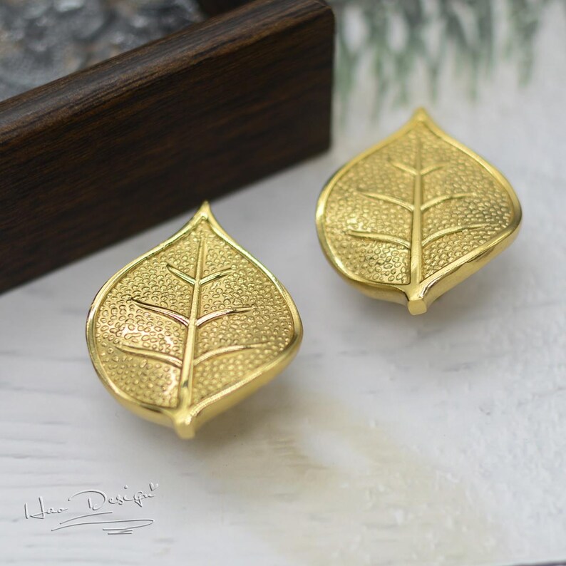 K&ouml;nnte beinhalten: Ein Paar goldene Ohrringe in Blattform. Die Bl&auml;tter haben eine strukturierte Oberfl&auml;che und ein detailliertes Adernmuster.