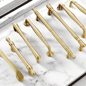 May include: A collection of gold-toned cabinet handles in various designs. The handles are arranged on a white and gray marble surface. The handles have different shapes, including curved, straight, and ornate styles.