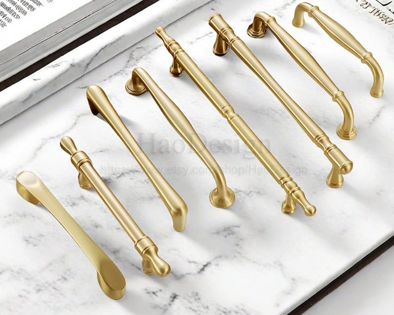May include: A collection of gold-toned cabinet handles in various designs. The handles are arranged on a white and gray marble surface. The handles have different shapes, including curved, straight, and ornate styles.