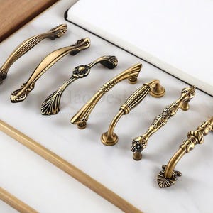 以下が含まれることがあります： さまざまな装飾的なデザインの、アンティーク真鍮のキャビネットプルとハンドルのコレクション。ハードウェアは、曲線的で詳細な形状で、暖かく、経年変化した仕上がりになっています。プルは白い表面に配置されています。