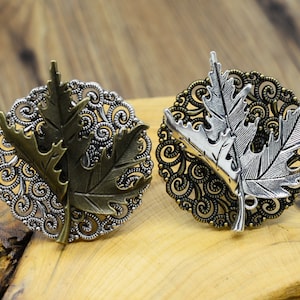 Op de afbeelding: Twee zilveren en bronzen broche's in de vorm van bladeren met ingewikkelde filigraanontwerpen. De broche's zijn geplaatst tegen een houten achtergrond.