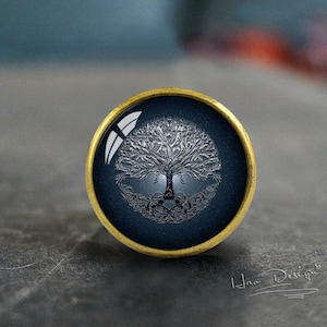 Könnte beinhalten: Ein runder Knopf in Goldton mit blauem Hintergrund und einem weißen Baum des Lebens-Design. Der Baum hat komplizierte Äste und Wurzeln.