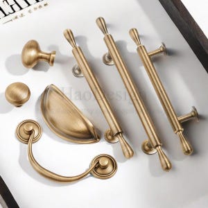 May include: An assortment of antique brass cabinet hardware, including knobs, pulls, and a cup pull. The hardware has a warm, aged finish and various shapes, suitable for drawers and doors. The hardware is arranged on a white surface.