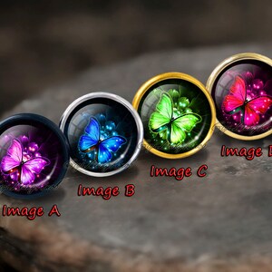 Puede incluir: Cuatro cabochones redondos de vidrio con engastes dorados, plateados y negros, cada uno con una mariposa de color diferente con alas iridiscentes. Las mariposas son rosa, verde, azul y morado.
