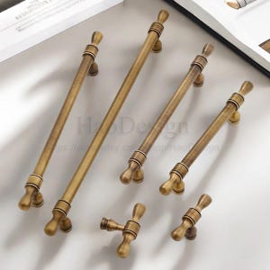 May include: A selection of antique brass cabinet pulls and knobs. The hardware has a warm, aged finish and decorative detailing. The pulls come in various sizes, with a T-shaped knob and a small knob. The text "HaoDesign" is visible.