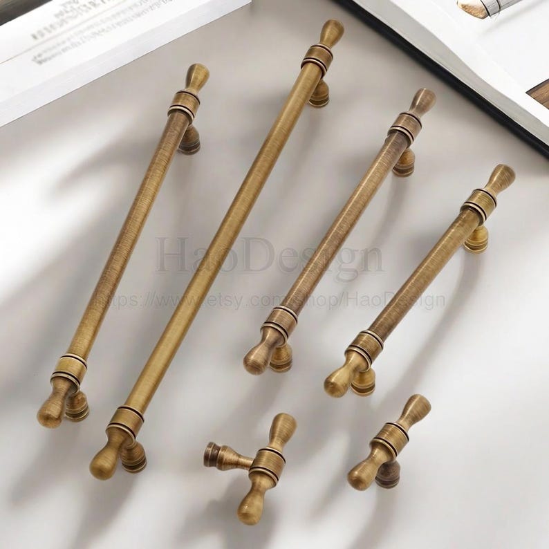 May include: A selection of antique brass cabinet pulls and knobs. The hardware has a warm, aged finish and decorative detailing. The pulls come in various sizes, with a T-shaped knob and a small knob. The text "HaoDesign" is visible.