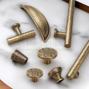 Könnte beinhalten: Sammlung antiker Messing-Schrankbeschläge, darunter Griffe und Knöpfe. Die Beschläge weisen eine gehämmerten Textur und eine warme, goldbraune Farbe auf. Verschiedene Formen und Größen werden gezeigt, geeignet für Möbel oder Schränke.