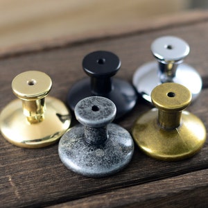 May include: Five different cabinet knobs in various finishes, including gold, brass, silver, black, and grey. The knobs are round and have a raised centre.
