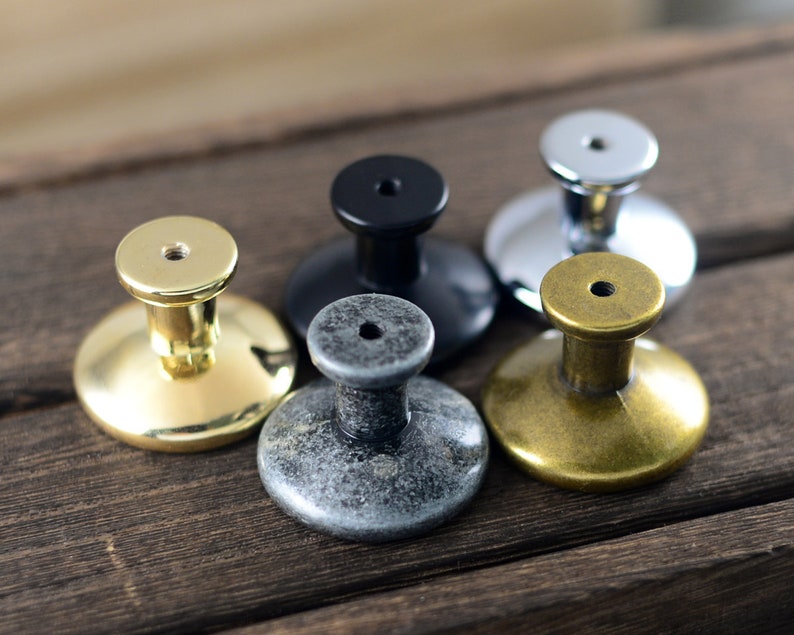 May include: Five different cabinet knobs in various finishes, including gold, brass, silver, black, and grey. The knobs are round and have a raised centre.