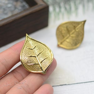 Może przedstawiać: Para złotych kolczyków w kształcie liści. Liście mają fakturowaną powierzchnię i szczegółowy wzór żyłek.