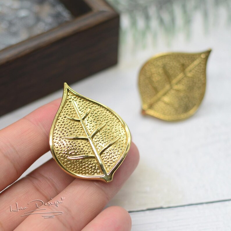 K&ouml;nnte beinhalten: Ein Paar goldene Ohrringe in Blattform. Die Bl&auml;tter haben eine strukturierte Oberfl&auml;che und ein detailliertes Adernmuster.